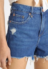 Shorts de denim en azul con un dobladillo deshilachado, detalles desgastados y herrajes de tono cobre. Cuenta con dos bolsillos frontales y un diseño de cintura alta.