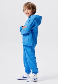Jongen staat met gekruiste armen, gekleed in een blauwe hoodie, bijpassende sweatpants en witte Nike sneakers met blauwe accenten.