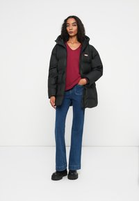 Crna puf jakna s kapuljačom, kopčanje na dugmad i bočni džepovi, nosi se preko burgundske dukserice i plavih traperica širom nogavica. Crna obuća.