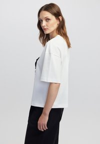 Witte T-shirt met korte mouwen en een ontspannen pasvorm, met zwarte tekst of versiering op de voorkant. Glad katoenen textuur, minimalistisch ontwerp.