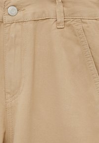 Close-up van beige broek met metalen knoop, riemlus en stiksels van de voorzak.