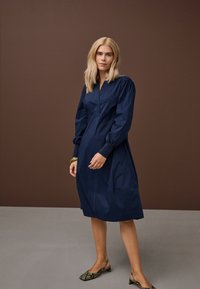 Ženska z blond lasmi, oblečena v mornarsko modro midi obleko z dolgimi rokavi in sandale z visoko peto iz kačjega usnja, stoji pred rjavim in sivim ozadjem.