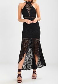 Robe noire en dentelle à col licou avec buste découpé, jupe fendue jusqu'au genou laissant apparaître une doublure florale blanche. Associée à des talons noirs à lanières.