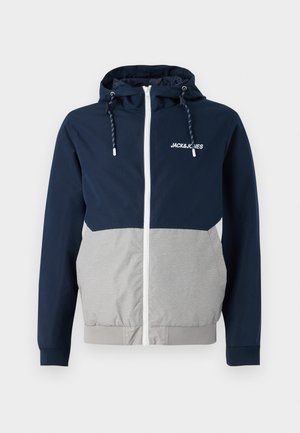 Chubasquero en navy y gris con cremallera frontal, capucha ajustable y logo "JACK&JONES"; presenta una textura suave y bloques de color contrastantes.