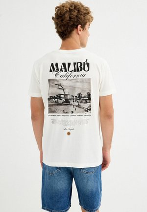 Hombre con cabello rizado vistiendo una camiseta blanca de Malibu California con un gráfico de playa en blanco y negro, combinado con pantalones cortos de mezclilla azules, de espaldas.