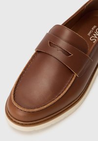 Mocasín de cuero marrón con acabado cosido, que presenta una tira y detalle de corte. Suela de goma color crema visible en la parte inferior. Textura suave.