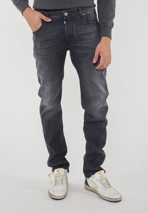 Persona che indossa jeans grigio scuro con una mano in tasca e sneakers bianche con suole beige e loghi arancioni.