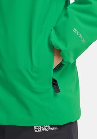 Grüne wasserdichte Jacke aus strukturiertem Stoff, mit einer Tasche auf der linken Seite und einem Logo am unteren Saum. "TEXAPORE"-Markierung auf dem Ärmel.