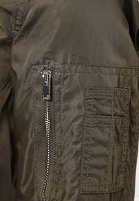 Olivově zelená bunda s hladkým, lesklým povrchem, která má stříbrný zip a detailem náprsní kapsy na levé straně.