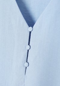 Blusa azzurra chiara con scollatura a V. Presenta tre bottoni rivestiti in tessuto e un morbido materiale in lino strutturato.