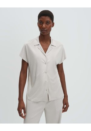 Femme debout portant une chemise boutonnée à manches courtes, beige clair, et un pantalon assorti, sur un fond clair uni.