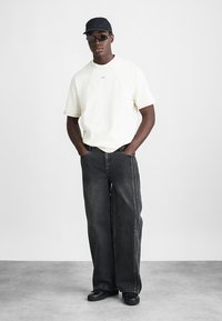 T-shirt de algodão branco com mangas curtas, combinada com jeans de ganga pretos folgados com costuras laterais e sapatos pretos. Um boné simples completa o visual.