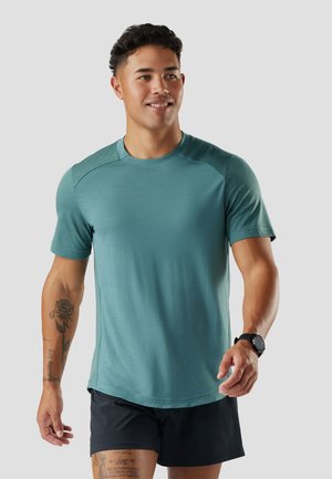 Ung mand med kort krøllet hår iført en teal atletisk skjorte, sorte shorts, sort smartwatch og synlige tatoveringer på venstre arm og ben.