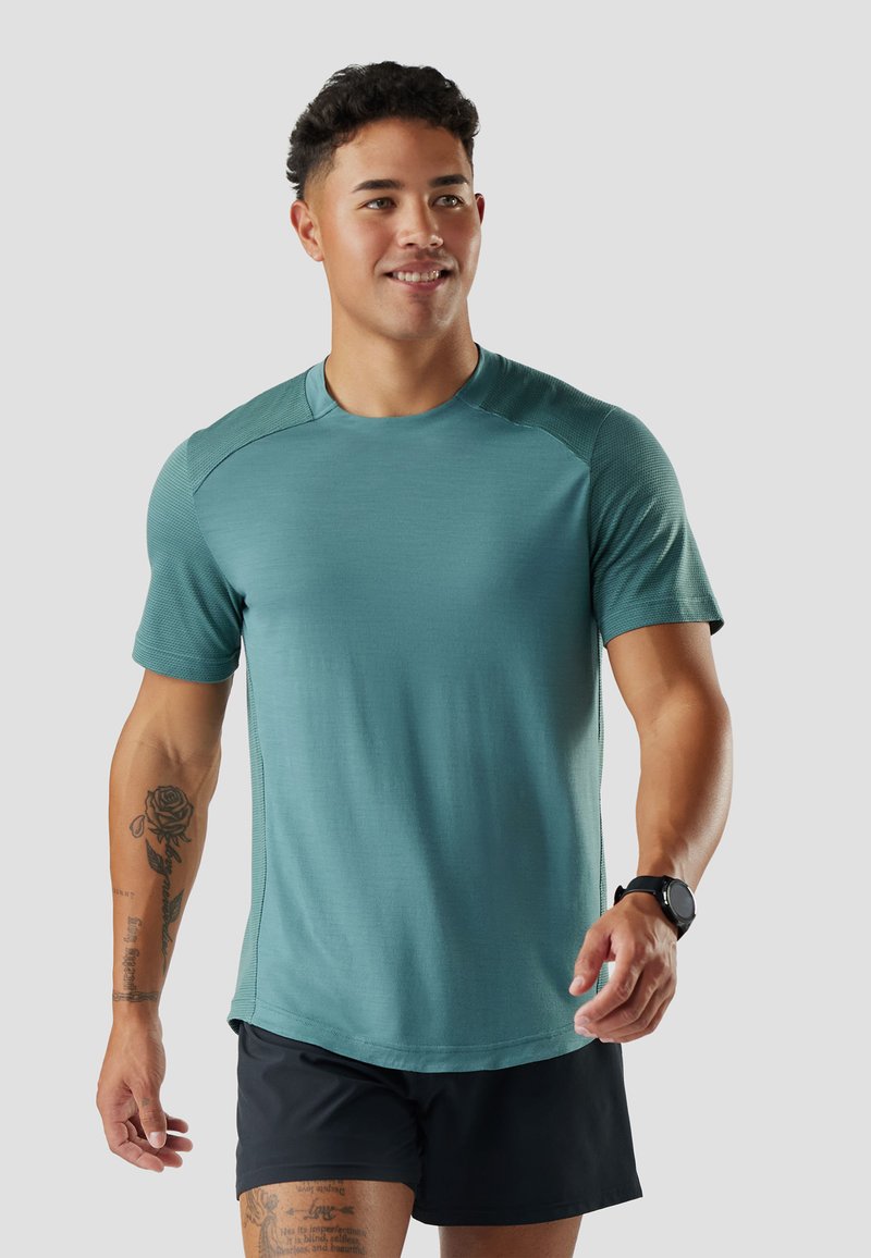 Giovane uomo con capelli corti e ricci che indossa una maglietta sportiva turchese, pantaloncini neri, smartwatch nero e tatuaggi visibili sul braccio sinistro e sulla gamba.