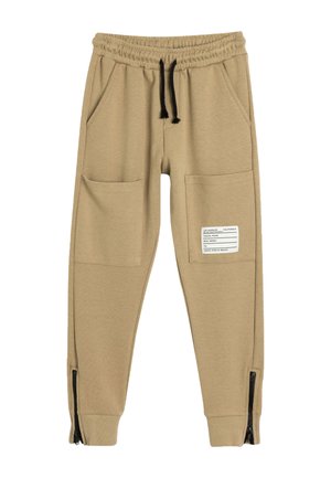 Pantalon de jogging beige en tissu doux avec une taille élastique, deux poches avant et des zippers aux poignets de cheville. Présente une étiquette sur le côté.