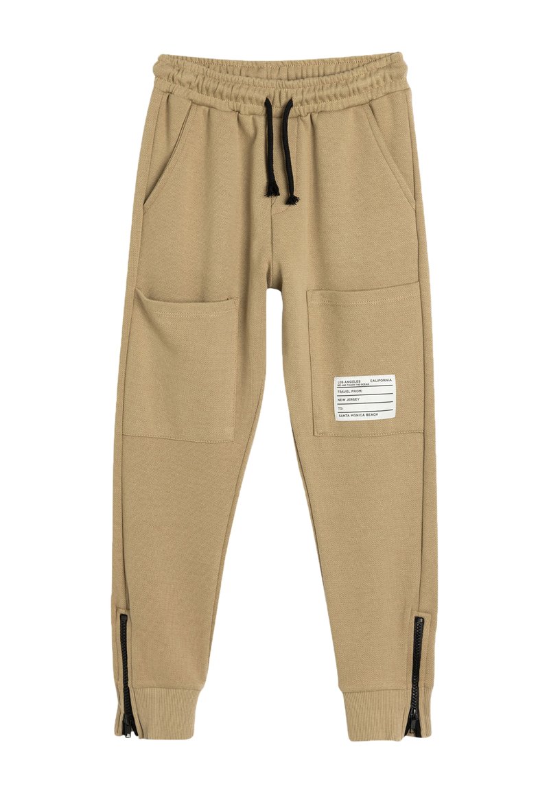 Beige joggerbukser i blødt stof med elastisk talje, to frontlommer og lynlåse ved ankelmanchetterne. Har et mærke på siden.