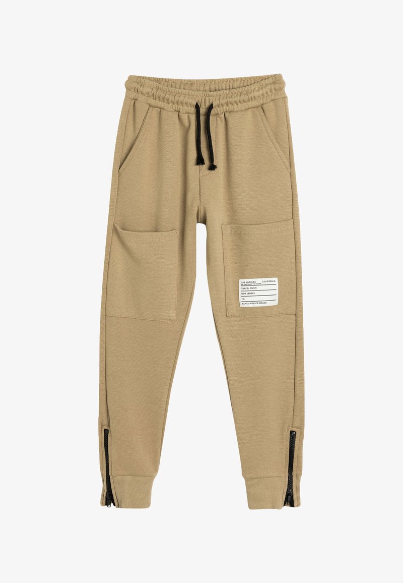 Beige jogger pants in soft fabric with an elastic waistband, two front pockets, and zippers at the ankle cuffs. Features a label on the side.