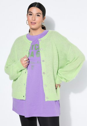 Nainen, jolla on tummat hiukset, pukeutuneena vaaleanvihreään napitettuun neulepuseroon violetin graafisen t-paidan ja mustien housujen päällä, seisoo valkoista taustaa vasten.