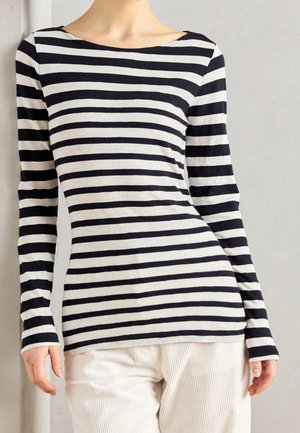 Personne portant une chemise à manches longues rayée noir et blanc et un pantalon de couleur claire sur un fond uni.
