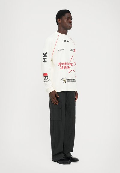 Man stands facing right wearing a white long-sleeve shirt with red and black graphic text and black wide cargo pants with pockets.