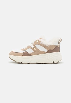 Zapatilla beige y blanca con suela gruesa, paneles de ante y cuero, cordones blancos, ojales dorados y forro de piel mullida alrededor del tobillo.