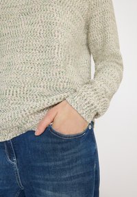 Personne portant un pull en tricot texturé avec la main dans la poche d'un jean en denim bleu, sur un fond uni.
