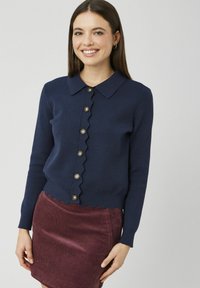 Jeune femme souriante, portant un cardigan bleu marine aux bords festonnés et à boutons, associé à une jupe en velours côtelé bordeaux.