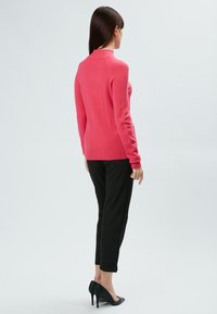 Rosa Strickpullover mit hohem Kragen und gerippten Bündchen, kombiniert mit schwarzen, maßgeschneiderten Hosen und schwarzen hohen Schuhen.
