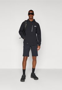 Black pullover hoodie with a front pocket and zip, paired with black shorts. Model wears dark sunglasses and a backpack, with black hiking shoes.