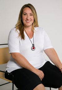 Top bianco a maniche corte e pantaloni corti neri. Grande pendente circolare con accentuazione rossa. Sfondo semplice, seduta su una sedia nera con sedile in legno.