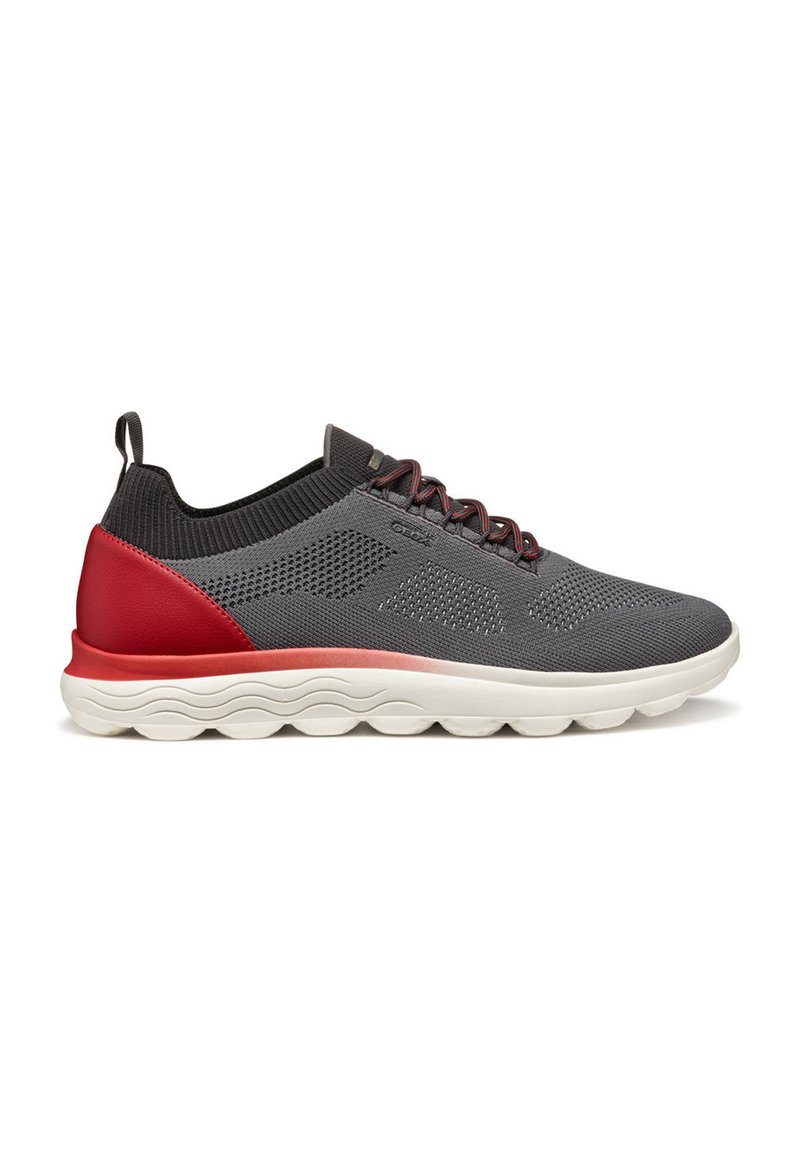 Scarpa sportiva in mesh grigia con un accento rosso sul tallone e una suola bianca texturizzata. Presenta lacci e un design con collo in maglia per un comfort aggiuntivo.