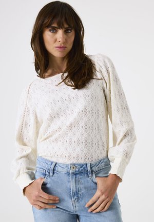Woman with brown hair wearing a white textured long-sleeve sweater and light blue jeans, posing with hands near pockets against white background.