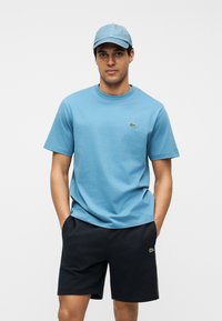 Homme portant une casquette bleu clair et un T-shirt avec un short noir, debout les mains dans les poches devant un fond blanc uni.