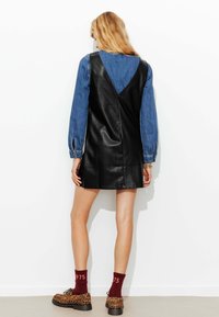 Robe en cuir noir avec un décolleté en V plongeant, superposée sur une chemise en denim bleu. Portée avec des chaussures à imprimé léopard et des chaussettes bordeaux avec des lettres.