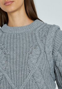 Grauer Strickpullover mit einem strukturierten Muster, das aus Zopfmuster-Designs und einem gerippten Rundhalsausschnitt besteht. Das Detail zeigt die Webart und die Nähte.