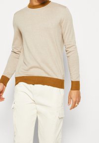 Pull beige en maille avec un col côtelé moutarde et un ourlet inférieur. Manches longues et coupe ajustée, porté avec un pantalon cargo de couleur claire.