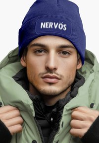 Blue knit beanie with a textured pattern and a patch that reads "NERVÖS," worn with a green puffer jacket against a neutral background.