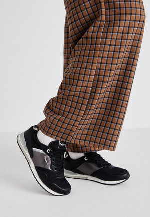 Sneaker nere e grigie con tomaia in pelle liscia, allacciate davanti, e suola bianca, abbinate a pantaloni a quadri marroni.