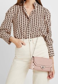 Světle růžová kožená kabelka se stříbrným řetízkovým popruhem, která má sklápěcí klopu, stříbrné kovové prvky a hřebíkované detaily.