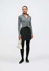 Pilkas ribuotas megztinis su užtrauktuku ir trumpintu kirpimu, juoda klostuota sijonas, juodos pėdkelnės ir plokšti batai, laikanti mažą baltą rankinę.