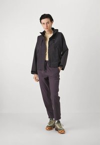 Άτομο με κοντά σκούρα μαλλιά φοράει μαύρο σακάκι, ανοιχτό μπεζ μπλούζα, σκούρες μοβ παντελόνες και πεζοπορικά παπούτσια σε πράσινο και πορτοκαλί, σε ουδέτερο φόντο.