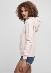 Világos rózsaszín kapucnis póló kenguru zsebbel, húzózsinórral ellátott kapucnival és bordázott mandzsettával. Farmer sorttal viselve, lazán állva.