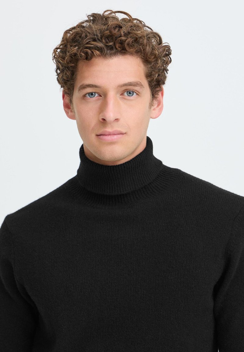 Jeune homme aux cheveux bruns bouclés et aux yeux bleus, portant un pull à col roulé noir, faisant face à un fond clair uni.