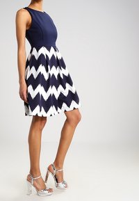 Mörkblå klänning med en figursydd liv och en utsvängd kjol med ett vitt zigzag-mönster. Silverfärgade högklackade sandaler kompletterar outfiten.