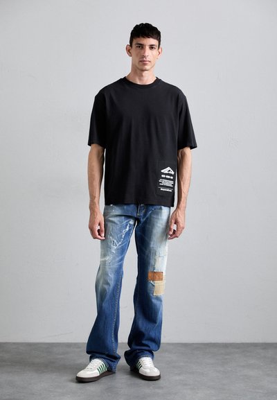 Black oversized t-shirt features a pocket with white graphic text. Worn with distressed blue jeans, displaying various patches and frayed edges.