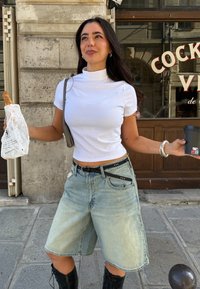 Witte korte mouw mock neck top, lichtblauwe denim bermuda shorts met een zwarte riem, houdt een snack in papier en een zwarte beker vast.