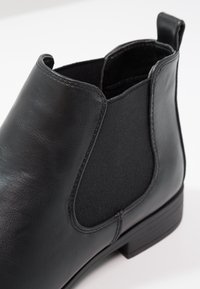 Bottine en cuir noir avec panneaux élastiques sur les côtés, talon plat et lignes épurées. Présente une finition en cuir texturé et des détails cousus.