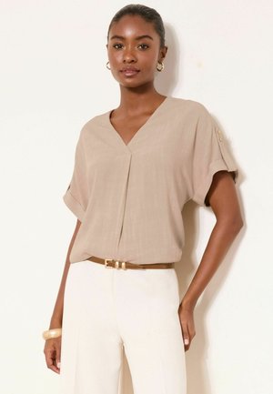 Kvinde iført beige V-hals kortærmet bluse med knappedetaljer på ærmerne, hvide bukser, brun bælte og guld øreringe.