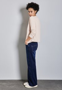 Světlý béžový prošívaný kabát s dlouhými rukávy, kombinovaný s tmavě modrými širokými džíny a bílými teniskami, umístěný na šedém pozadí.