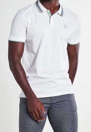 Man draagt een witte korte mouw polo met groen afgewerkte kraag en mouwen, gecombineerd met nauwsluitende grijze broek met textuur.